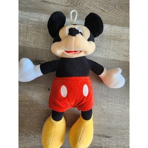 May include: A plush Mickey Mouse toy with black ears, a black body, red shorts with white circles, and yellow shoes. The toy has a friendly expression with a big smile.
