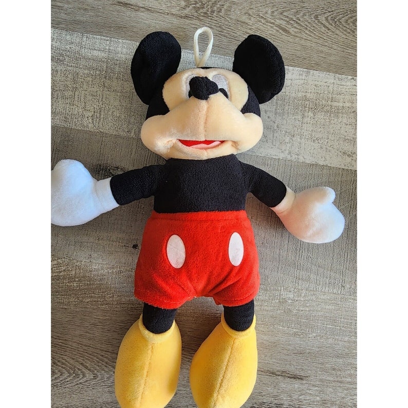 May include: A plush Mickey Mouse toy with black ears, a black body, red shorts with white circles, and yellow shoes. The toy has a friendly expression with a big smile.