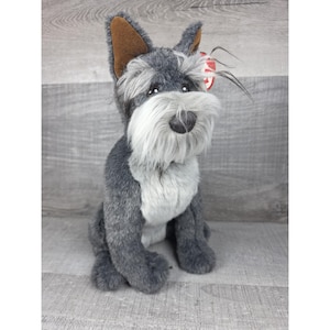 Op de afbeelding: Een pluche Schnauzer hond met grijze vacht, een witte baard en bruine oren. Het knuffeldier zit rechtop, met een rood label aan zijn oor. De hond heeft een zwarte neus en kleine, donkere ogen.
