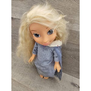 May include: A blonde doll wearing a blue dress with a white fur trim around the neck and a blue cape. The doll has blue eyes and a smiling face.