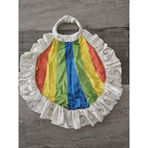 Può includere: Un mantello arcobaleno con un bordo di volant bianchi e un anello bianco in cima. Il mantello ha un motivo a strisce rosse, gialle, verdi, blu e viola con pois bianchi.