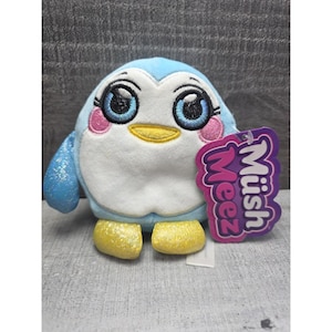 Op de afbeelding: Een pluche pinguïn speelgoed met een blauw lichaam, witte buik en gele voeten. De pinguïn heeft grote blauwe ogen, roze wangen en een gele snavel. Een paars label met de tekst "Müsh Meez" is bevestigd.