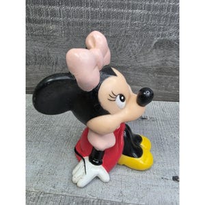 May include: A ceramic figurine of Minnie Mouse, featuring a black head and ears, a light pink bow, and a red dress. Her shoes are yellow, and her gloves are white. The figurine is in a seated position.