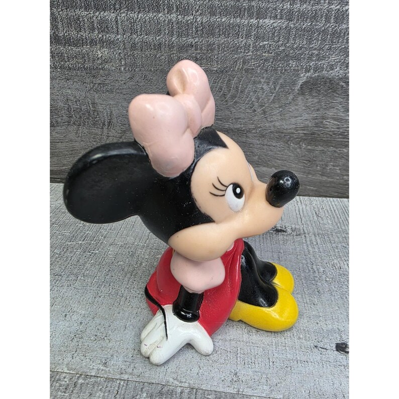 May include: A ceramic figurine of Minnie Mouse, featuring a black head and ears, a light pink bow, and a red dress. Her shoes are yellow, and her gloves are white. The figurine is in a seated position.
