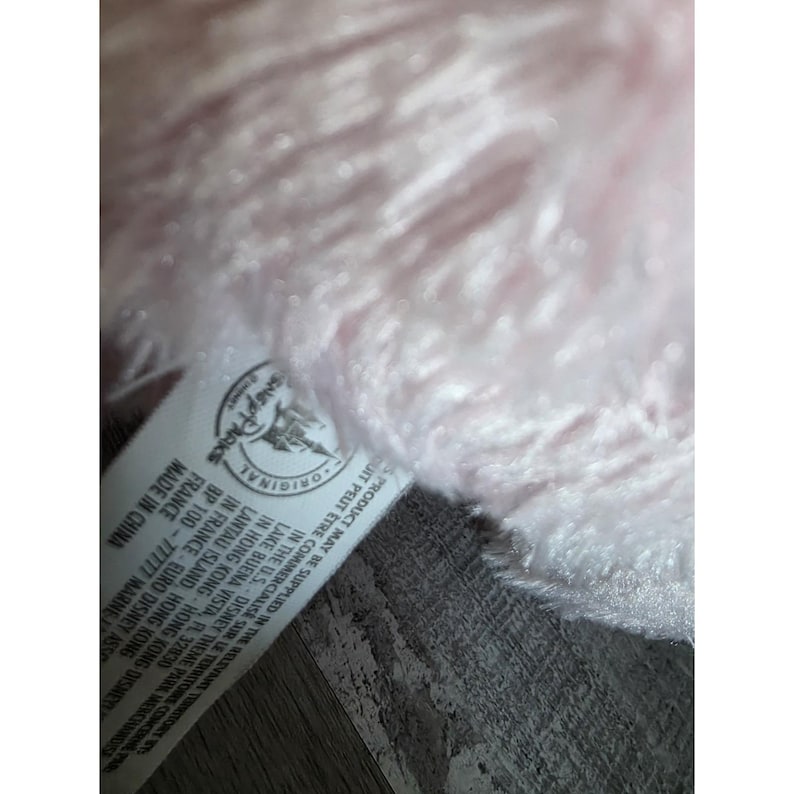 May include: Close-up of a soft, pink plush toy. A tag with the text "Disney Parks Original" and other text is visible. The toy's fur is fluffy and textured, with a light pink hue.