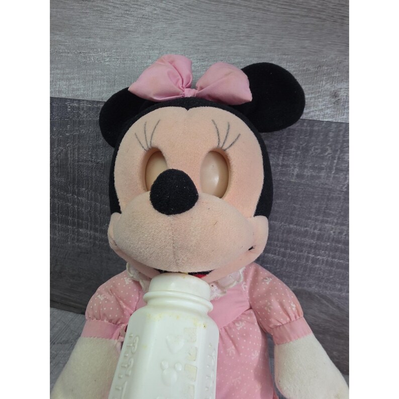 Puede incluir: Una mu&ntilde;eca de peluche vintage de Minnie Mouse con un vestido rosa con lunares blancos y un lazo a juego. La mu&ntilde;eca sostiene un biber&oacute;n blanco. La mu&ntilde;eca tiene orejas negras y una nariz negra.