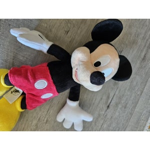 Puede incluir: Un juguete de peluche de Mickey Mouse con orejas negras, una camisa roja y blanca y zapatos amarillos. El juguete est&aacute; acostado sobre una superficie de madera.