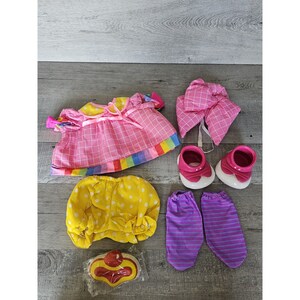 May include: A collection of doll clothes, including a pink checkered dress with a rainbow trim, a yellow polka-dot diaper cover, and a pink bonnet. Also included are pink shoes, purple striped socks, and a clown nose.