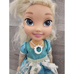 May include: A blonde doll wearing a blue and white dress with a snowflake design. She has a pearl necklace with a blue gem pendant.