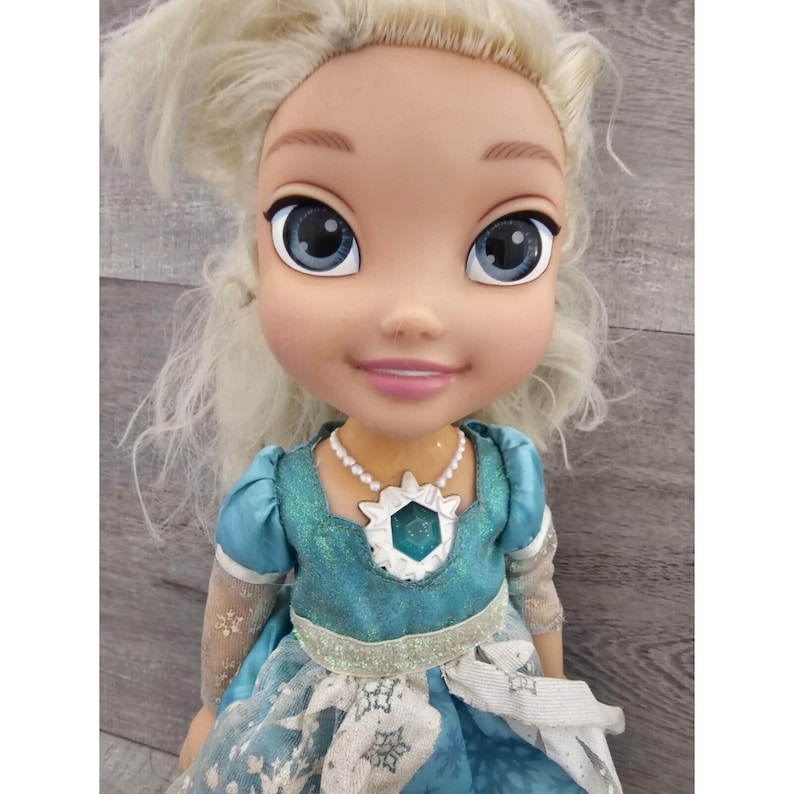 May include: A blonde doll wearing a blue and white dress with a snowflake design. She has a pearl necklace with a blue gem pendant.