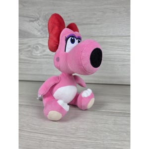 May include: A pink plush toy character with a large red bow on its head. The figure has a rounded snout with a black nose, white belly patch, and white accents on its feet. It features large eyes with purple eyelids and eyelashes, standing upright.