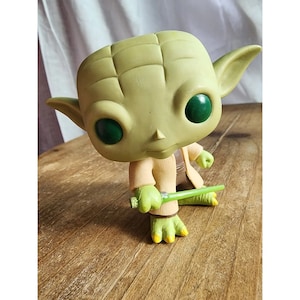 Op de afbeelding: Een groene en bruine Yoda Pop-figuur die een groen lichtzwaard vasthoudt. De figuur heeft grote groene ogen en een bruine robe.