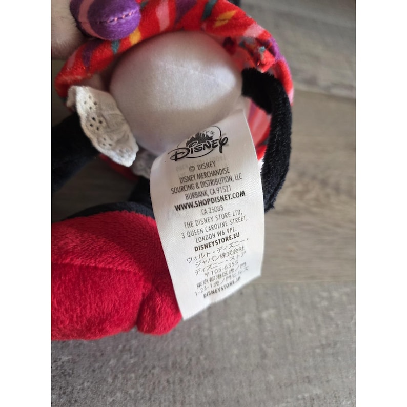May include: A plush toy featuring a white face, black ears, and red and black clothing. The toy has a red and patterned hat. A white tag with the Disney logo and website address is visible.
