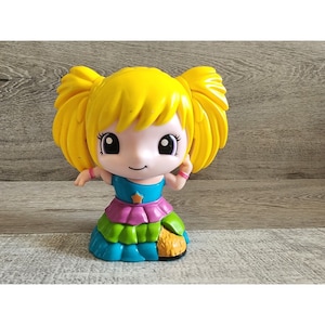 May include: A toy girl with yellow hair, wearing a blue, green and pink dress. She has a star on her dress and is smiling.