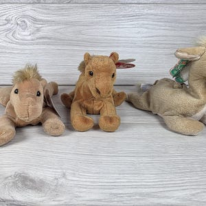 May include: Three tan plush camel toys in a lying position. The left camel has a light tan mane, the center camel has a darker tan coat with a red heart-shaped tag, and the right camel features a patterned green and white fabric accent on its back.