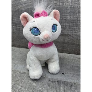 May include: A white plush cat with blue eyes, a pink nose, and a pink bow on its head. The cat has a pink scarf around its neck and is sitting upright. The plush toy is made of soft, fluffy material.