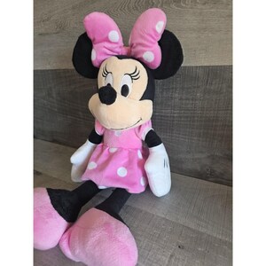 May include: A plush Minnie Mouse doll. The doll has black ears, a black nose, and a pink bow with white polka dots. She wears a pink dress with white polka dots, white gloves, and pink shoes. The doll is sitting on a wooden surface.