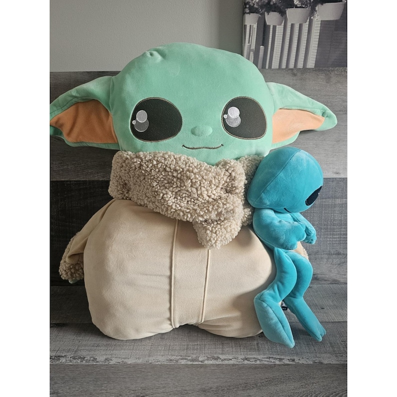 May include: A plush toy of a green alien character with large eyes and a brown scarf. The alien is holding a smaller blue alien plush toy.