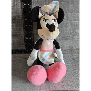K&ouml;nnte beinhalten: Ein Pl&uuml;schtier von Minnie Mouse, das ein rosa Kleid mit einem karierten Rock und einer karierten Schleife tr&auml;gt. Das Spielzeug hat schwarze Augen, eine schwarze Nase und ein rosafarbenes L&auml;cheln.
