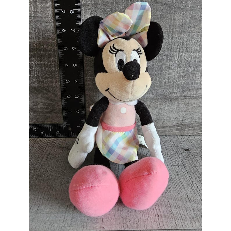 K&ouml;nnte beinhalten: Ein Pl&uuml;schtier von Minnie Mouse, das ein rosa Kleid mit einem karierten Rock und einer karierten Schleife tr&auml;gt. Das Spielzeug hat schwarze Augen, eine schwarze Nase und ein rosafarbenes L&auml;cheln.