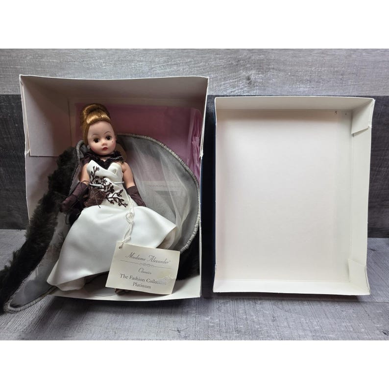 May include: A Madame Alexander doll in a white dress with brown detailing, presented in its original box. The doll has a blonde updo and is wearing brown gloves. The box is open, showing the doll and its accessories.