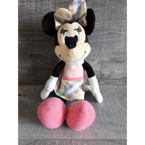 K&ouml;nnte beinhalten: Ein Pl&uuml;schtier von Minnie Mouse, das ein rosa Kleid mit Karomuster und eine passende Karoblume tr&auml;gt. Das Spielzeug hat schwarze Augen, eine schwarze Nase und ein rosafarbenes L&auml;cheln.
