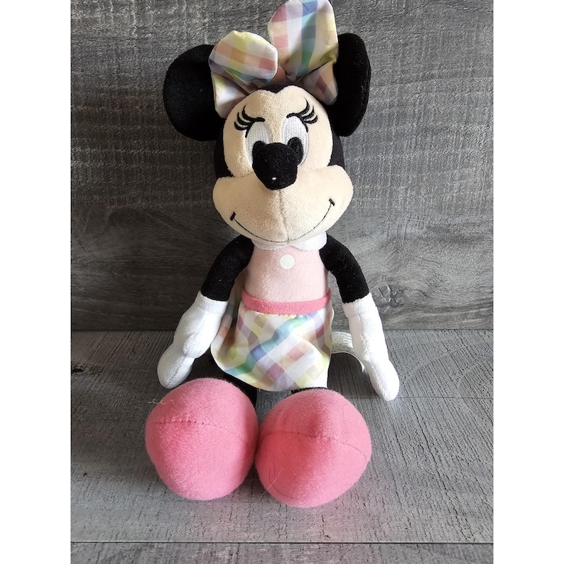K&ouml;nnte beinhalten: Ein Pl&uuml;schtier von Minnie Mouse, das ein rosa Kleid mit Karomuster und eine passende Karoblume tr&auml;gt. Das Spielzeug hat schwarze Augen, eine schwarze Nase und ein rosafarbenes L&auml;cheln.