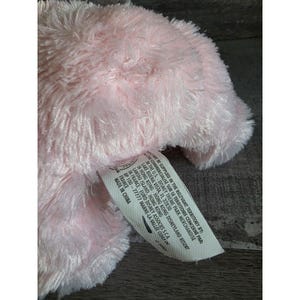 May include: A close-up of a soft, pink plush toy. The plush has a fluffy texture and is partially visible. A white tag with black text is attached to the toy. The tag includes text in English and French.