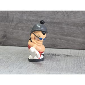 May include: A small, cartoonish figurine of a character with a tan body, red shorts, and a gray helmet. The character wears blue sunglasses and has a stern expression. The figurine is standing on a white and black base.