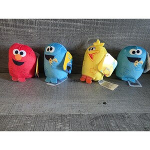 Op de afbeelding: Vier pluche speelgoed van Sesame Street-personages: Elmo in rood, twee Cookie Monsters in blauw en Big Bird in geel. Elk pluche heeft een label. Het speelgoed ligt op een grijs houten oppervlak.