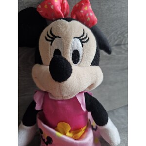 May include: A plush toy of Minnie Mouse wearing a pink dress with a yellow bow and a red bow with white polka dots in her hair.