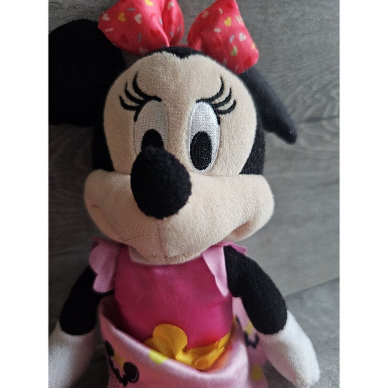 May include: A plush toy of Minnie Mouse wearing a pink dress with a yellow bow and a red bow with white polka dots in her hair.