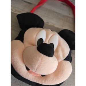 May include: A plush Mickey Mouse face bag with a red strap. The bag features Mickey's iconic black ears, a black nose, and large white eyes with black pupils. The face is a soft, light pink color, and the mouth is a darker pink.