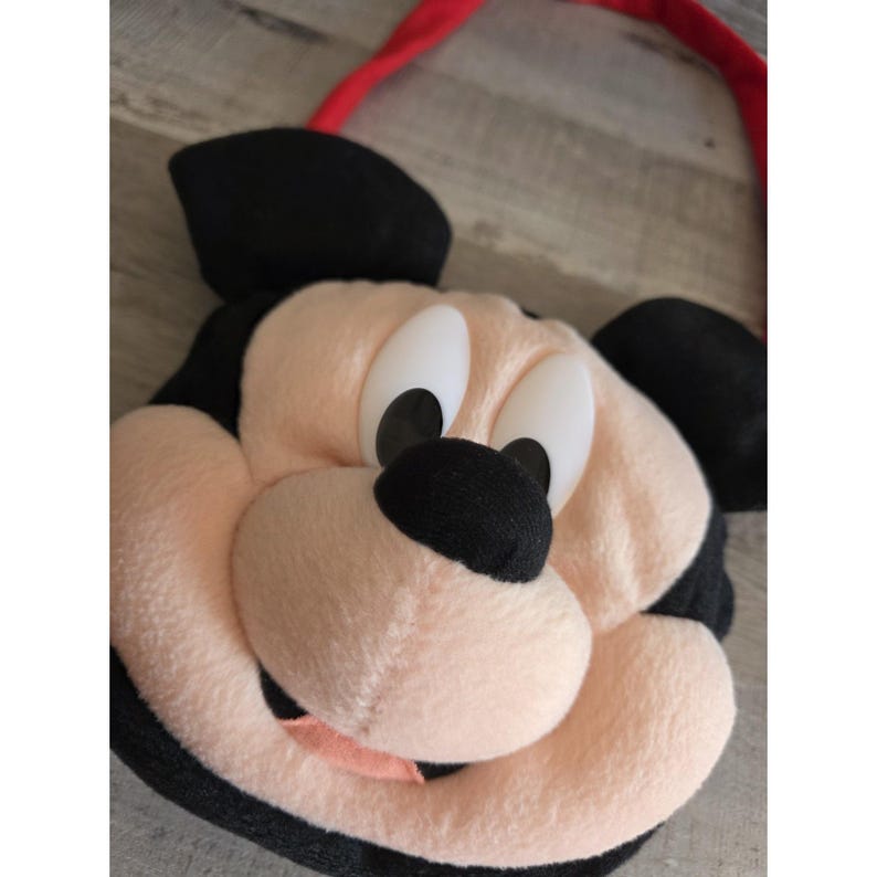 May include: A plush Mickey Mouse face bag with a red strap. The bag features Mickey's iconic black ears, a black nose, and large white eyes with black pupils. The face is a soft, light pink color, and the mouth is a darker pink.