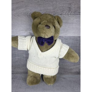 May include: A plush teddy bear with olive-brown fur, wearing a cream-colored V-neck sweater and a dark purple velvet bow tie. The bear has a dark brown nose and is standing with its arms outstretched.