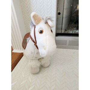May include: White plush toy horse with brown saddle and a brown bridle. The horse has a white mane and tail and brown eyes.