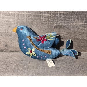 May include: A blue plush bird ornament with embroidered floral details. The bird has a yellow beak, a brown branch, and pink and green floral accents. The bird's tail feathers are a darker shade of blue.
