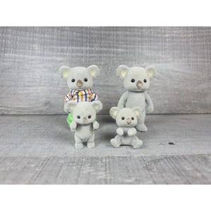 May include: Four gray koala figurines. Two are standing, one is sitting, and one is wearing a plaid shirt. The koalas have small black eyes and tan ears. The background is a gray wooden surface.