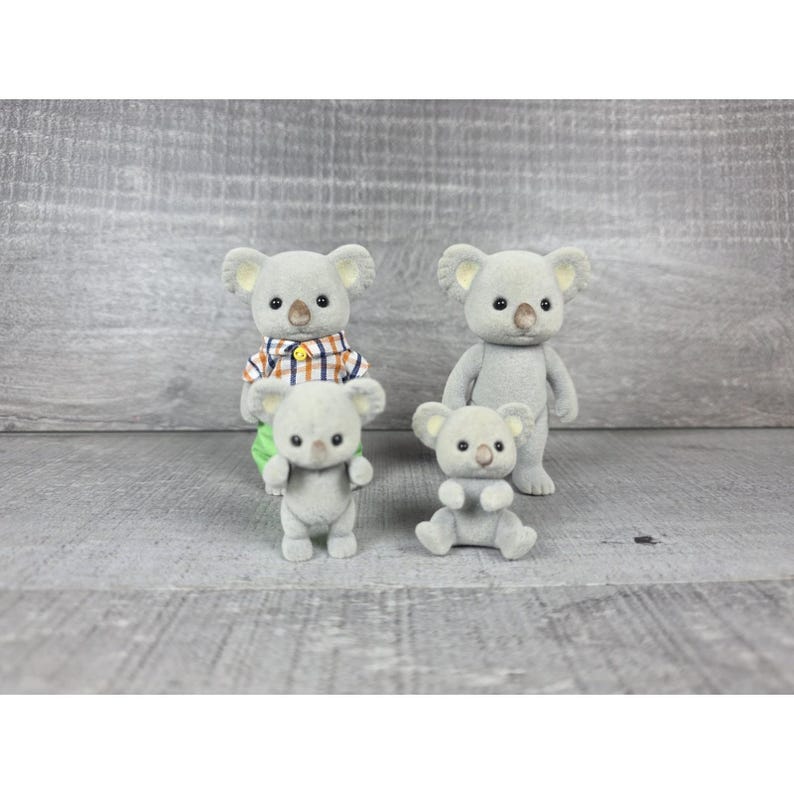 May include: Four gray koala figurines. Two are standing, one is sitting, and one is wearing a plaid shirt. The koalas have small black eyes and tan ears. The background is a gray wooden surface.