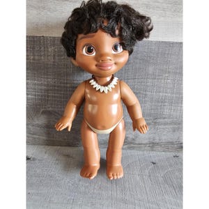 Pu&ograve; includere: Una bambola di pelle marrone con capelli scuri e ricci, che indossa una collana di conchiglie bianche e un bikini marrone. La bambola ha un viso sorridente con occhi marroni e sta in piedi su una superficie di legno.