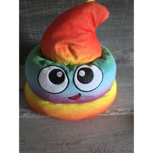 Op de afbeelding: Een pluche speelgoed in de vorm van een lachende regenboogkleurige poep-emoji. De bovenkant is een verloop van rood, oranje en geel, terwijl de basis een mix is van blauw, groen en geel. Het heeft grote cartoonogen.