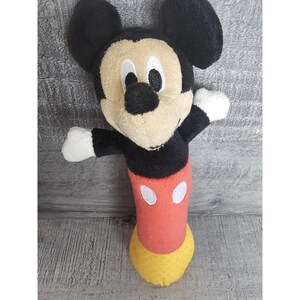 May include: A plush Mickey Mouse toy. It has a black head and ears, a tan face, and a black nose. The body is red with two white oval buttons, and the feet are yellow. The toy has white gloves and is in a standing position.