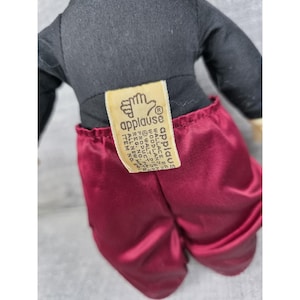 May include: A plush doll with a black top and burgundy satin pants. The doll has a tag that reads "applause" and "Walt Disney Product". The doll's arms and legs are not visible in the image.