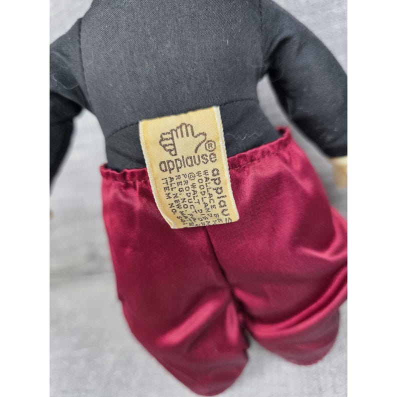 May include: A plush doll with a black top and burgundy satin pants. The doll has a tag that reads "applause" and "Walt Disney Product". The doll's arms and legs are not visible in the image.