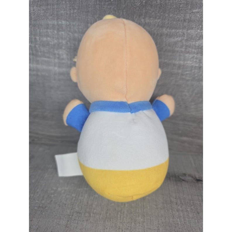 May include: A soft toy doll with a light tan head and a body wearing a white shirt with blue trim and sleeves, and a yellow bottom. The doll has small arms and a tag attached to the bottom.