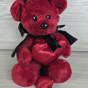 Op de afbeelding: Een pluche teddybeer met donkerrode vacht, zwarte hoorns en een witte haarlok op het hoofd. De beer draagt een zwarte cape die met een lint om de nek is gestrikt en houdt een glanzend rood satijnen hart vast. Een kleine hartvormige staart is zichtbaar.