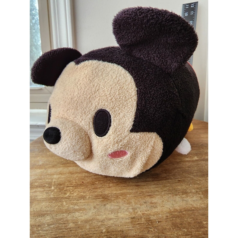 May include: A plush Mickey Mouse toy with brown ears, a black nose, and a beige face. The toy has large black eyes and pink cheeks.