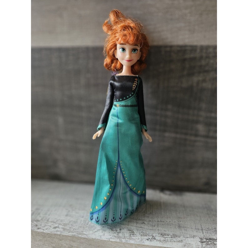May include: A doll with red hair wearing a black and teal dress with gold accents. The doll has a detailed face with brown eyes and a friendly expression.