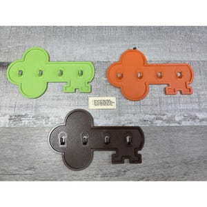 May include: Three key-shaped wall organizers in green, orange, and brown. Each key has four keyhole-shaped hooks. A small white card with instructions is visible. The organizers are made of plastic and are mounted on a gray wooden surface.