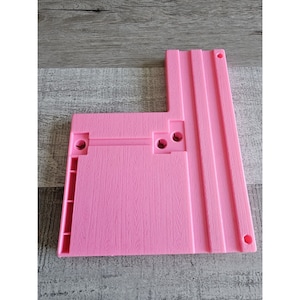 May include: Pink plastic track piece for a toy train set. The piece has a straight section and a curved section, with two holes on the straight section for connecting to other pieces.